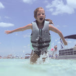 Child splashing in water while wearing the BOTE Youth Foam PFD Vest in gray, teal, and black.